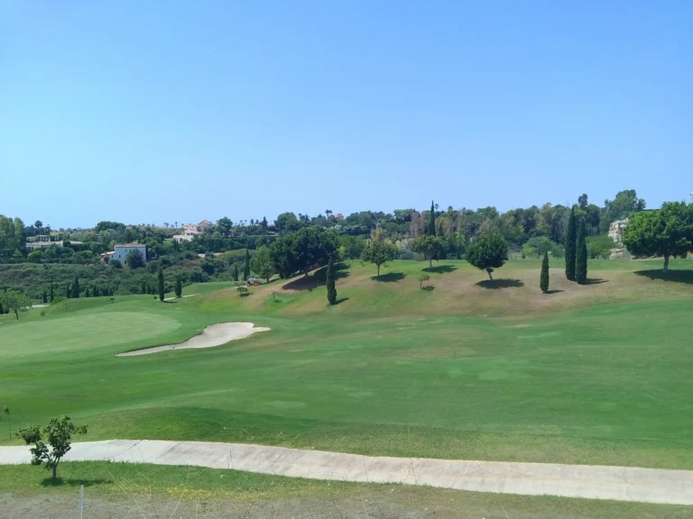 Golf green with a sand bunker and a cart path, with trees and houses on the ridge behind